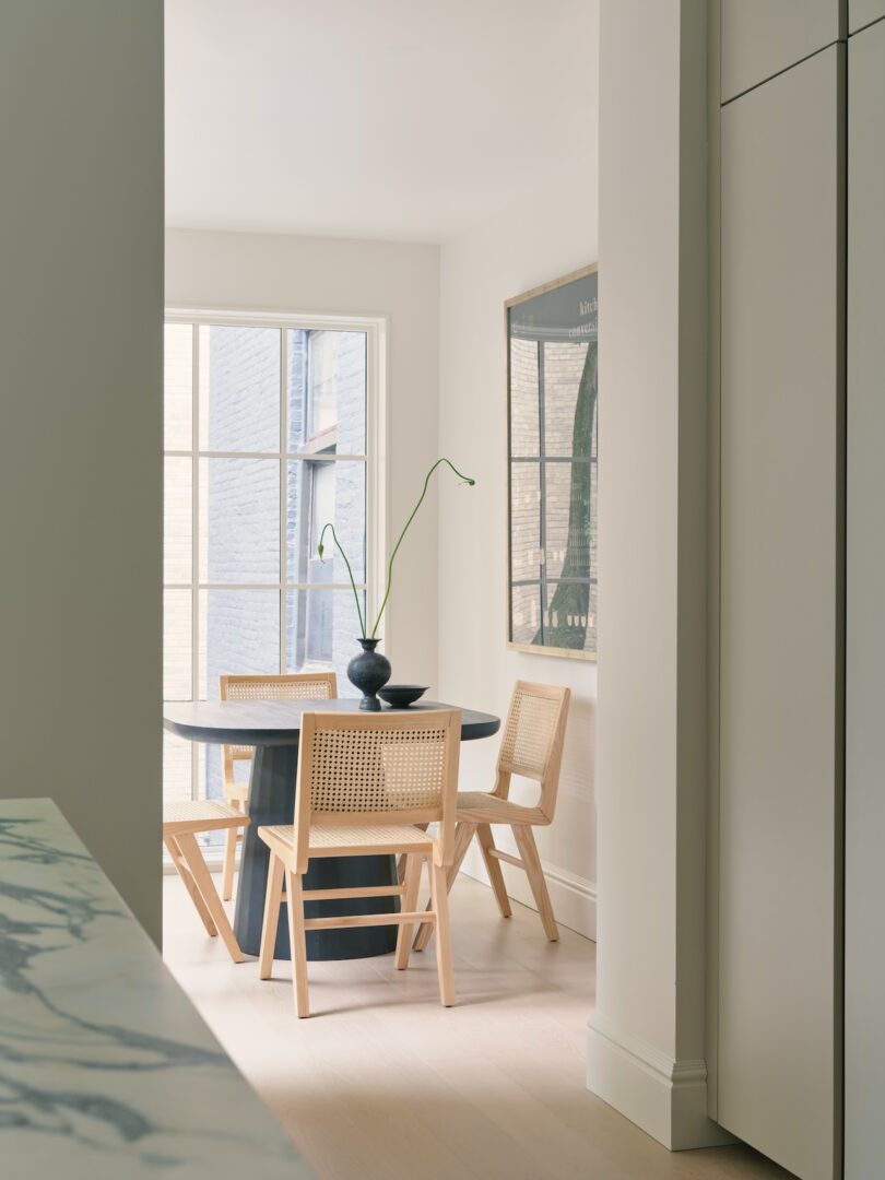 A minimalist dining area with a round black table, four wooden chairs, a vase with tall stems, large window, and wall art.