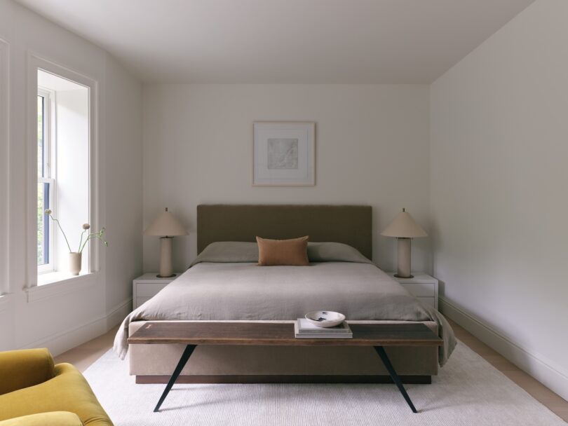 A minimalist bedroom with a neatly made bed, two bedside tables with lamps, a framed picture above the bed, a bench at the foot, and a window letting in natural light.