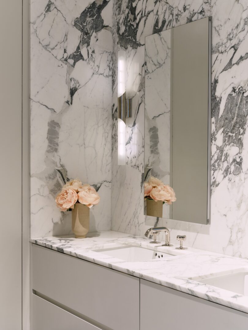 Modern bathroom with white marble walls and countertop, built-in sink, rectangular mirror, wall light, and a vase of light pink flowers on the counter.