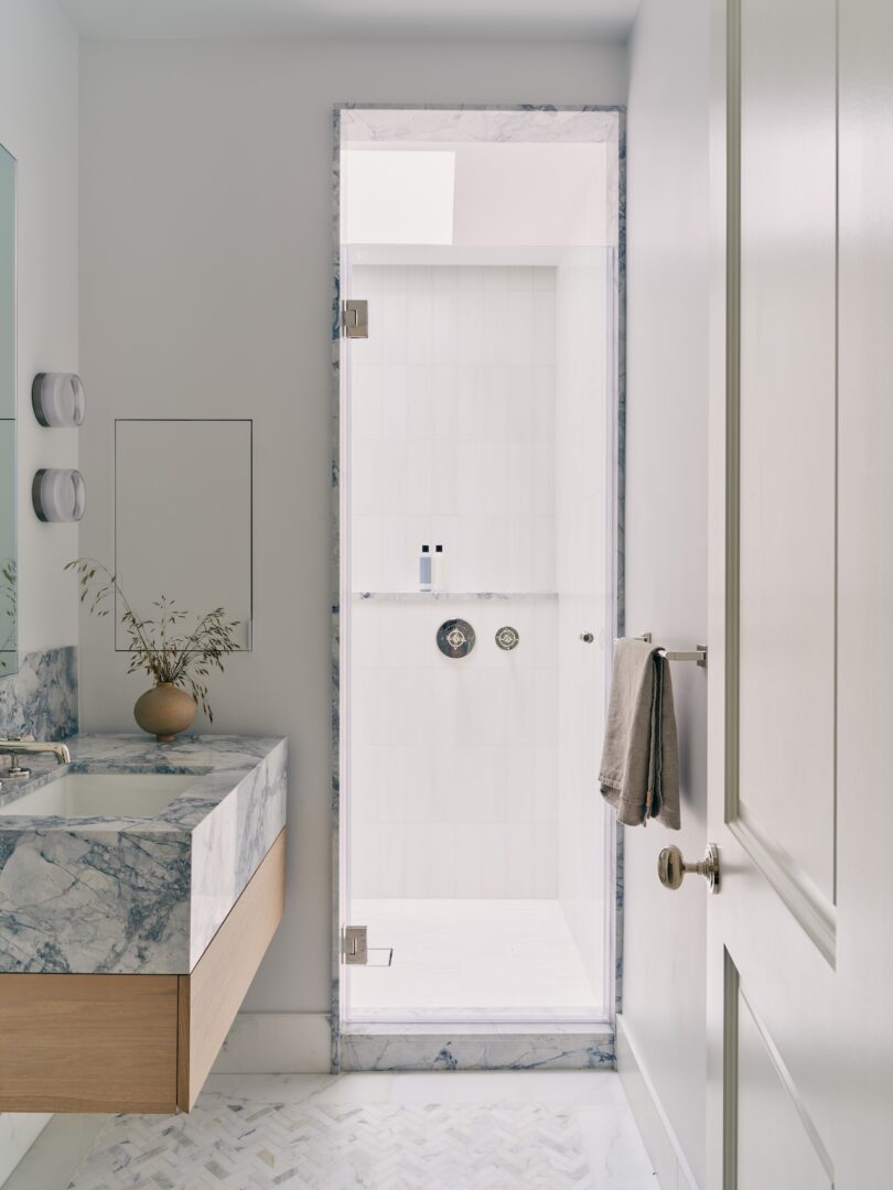 Modern bathroom with marble surfaces, floating vanity, a glass shower door, wall-mounted towel, and a small vase with branches on the counter.