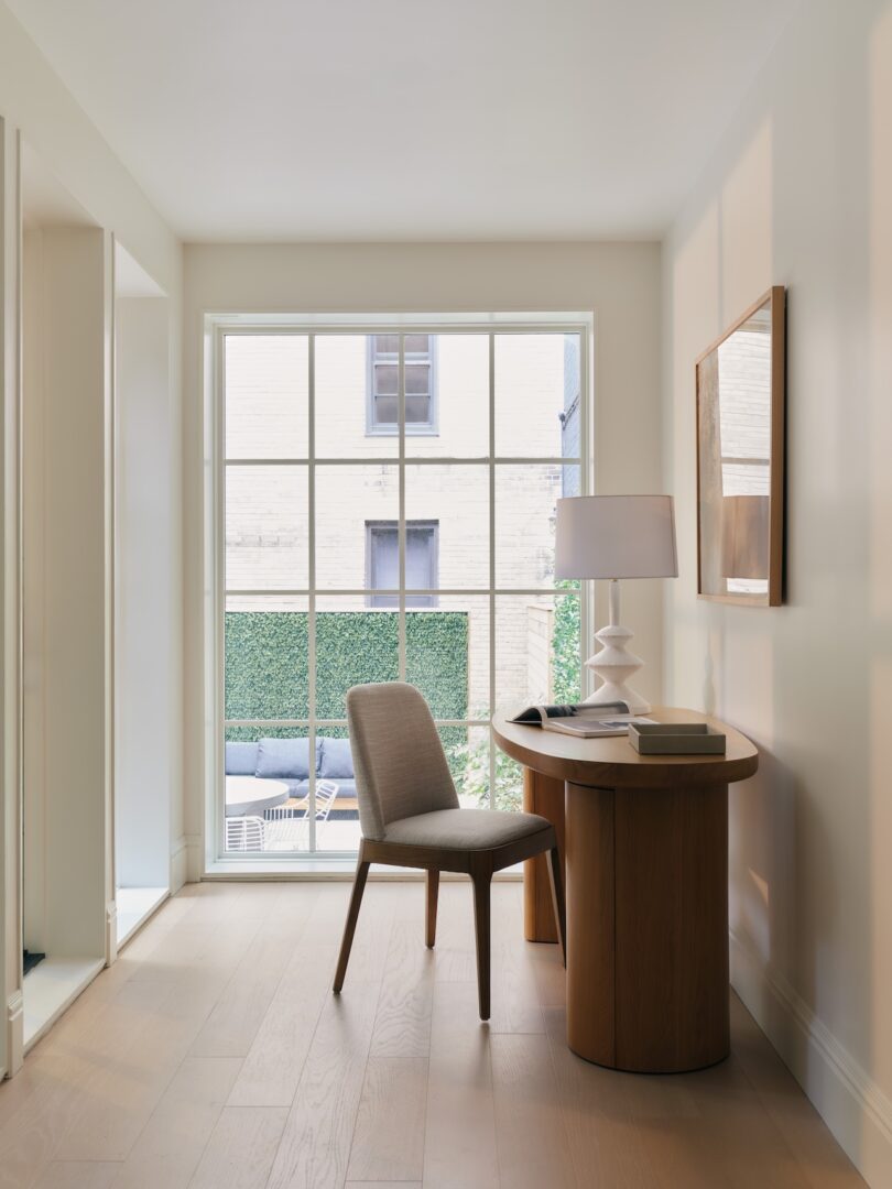 A small workspace with a wooden desk, beige chair, white table lamp, and books sits by a large window overlooking a courtyard with greenery.