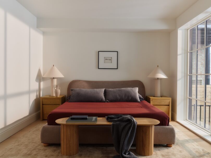 A neatly made bed with a red blanket, gray pillows, and a wooden bench sits between two nightstands with lamps in a bright, modern bedroom with large windows.