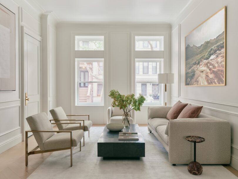 A modern living room with neutral tones features two armchairs, a sofa, a black coffee table, a small side table, a vase with greenery, and large windows letting in natural light.