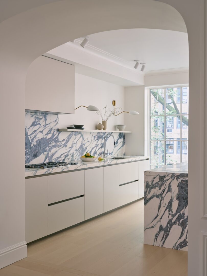 Modern kitchen with white cabinets, blue-veined marble countertops and backsplash, open shelves, light wood floor, and large window with natural light.