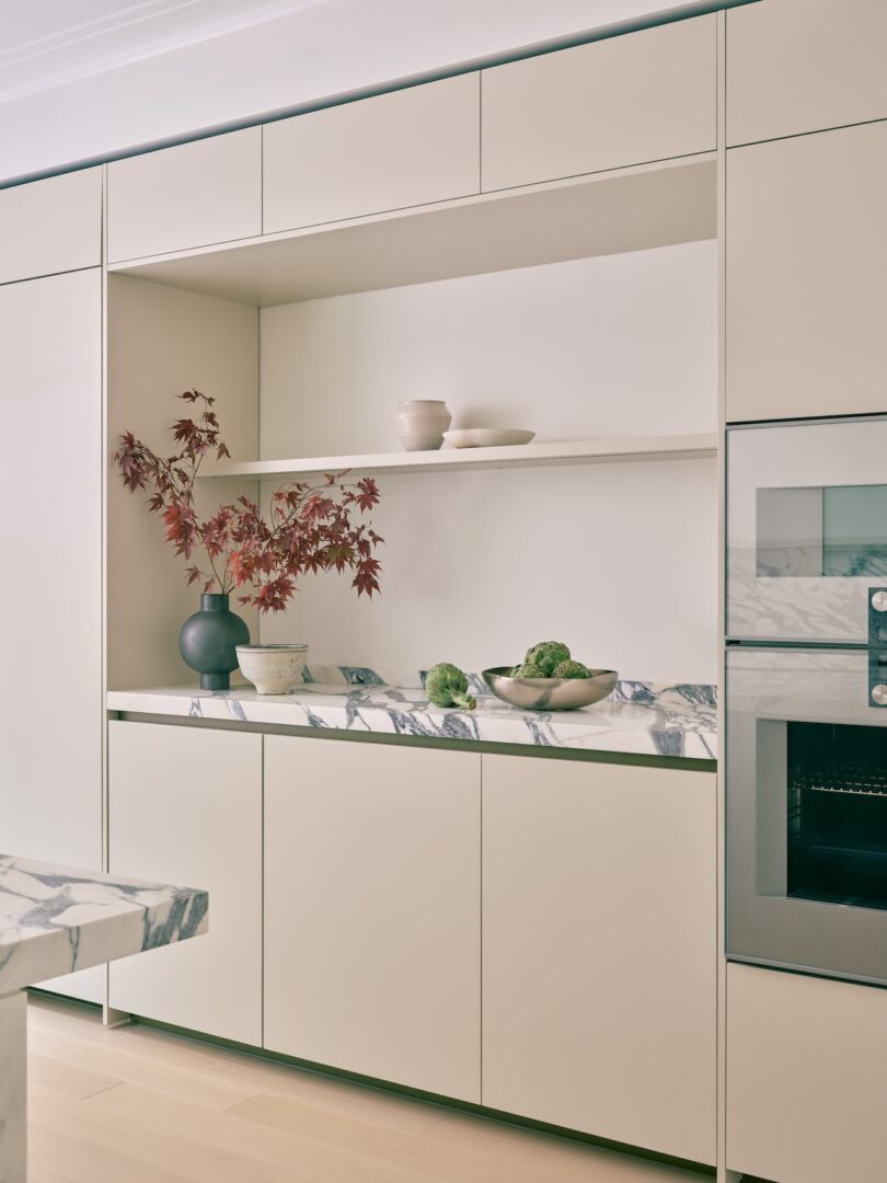 Minimalist kitchen with light beige cabinets, marble countertop, open shelving with bowls and a vase holding red leaves, and a built-in oven on the right.