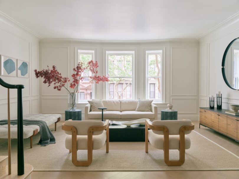 A modern living room with a cream sofa, two wooden armchairs, a wooden sideboard, a round mirror, large windows, and a coffee table with a vase of red branches.