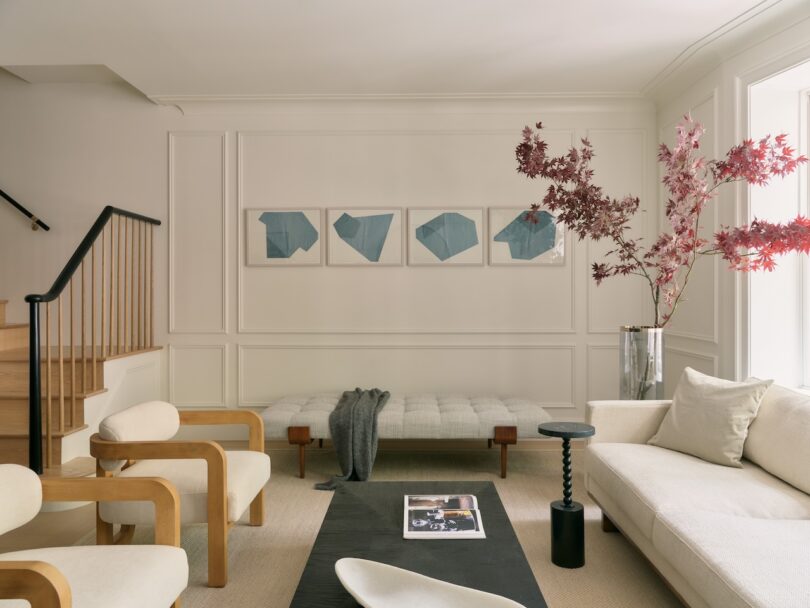 Modern living room with light-toned furniture, five blue abstract artworks on the wall, a glass vase with red leaves, and natural light from a large window.