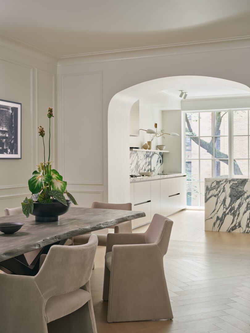Modern dining area with a stone table and six beige chairs, adjacent to a white kitchen with marble surfaces and large window, featuring minimalist decor and neutral tones.