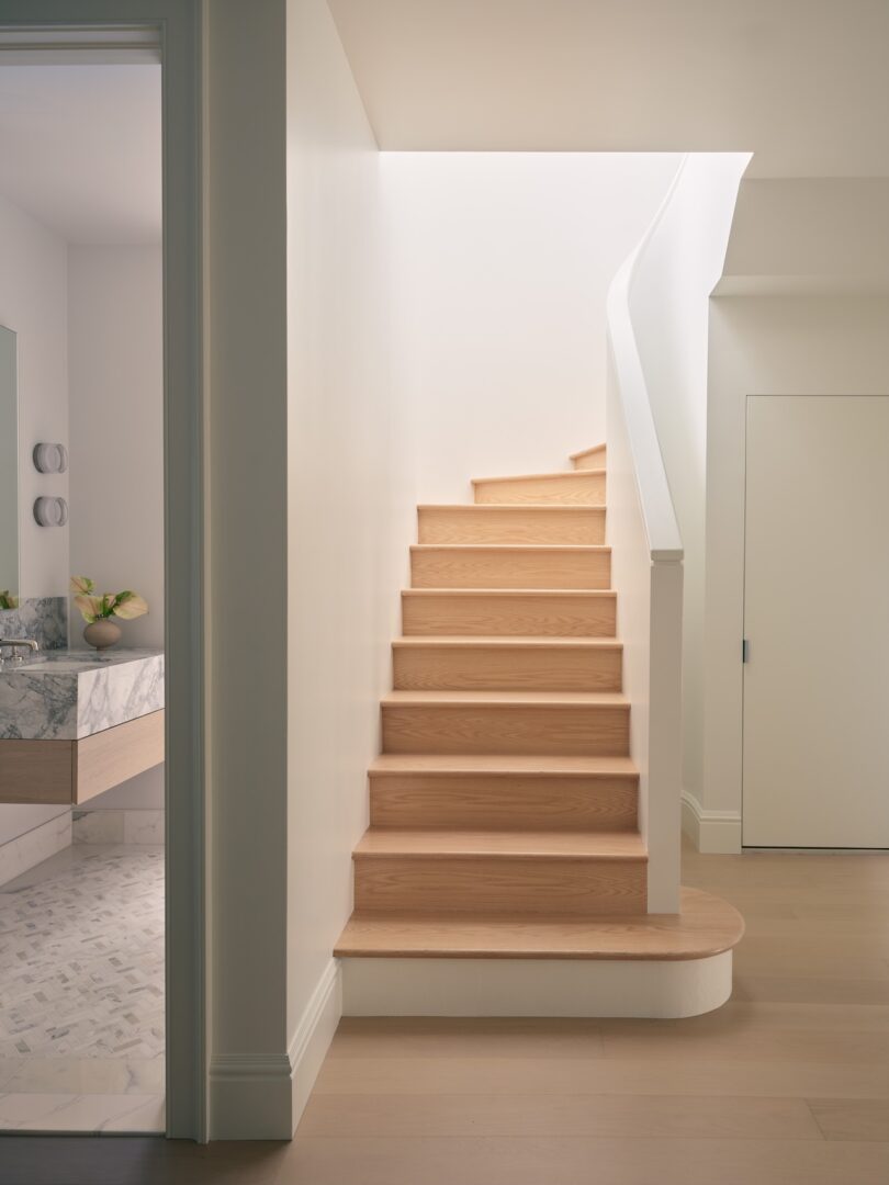 A light wood staircase with white walls curves to the right, next to a closed door and a bathroom with marble surfaces visible on the left.