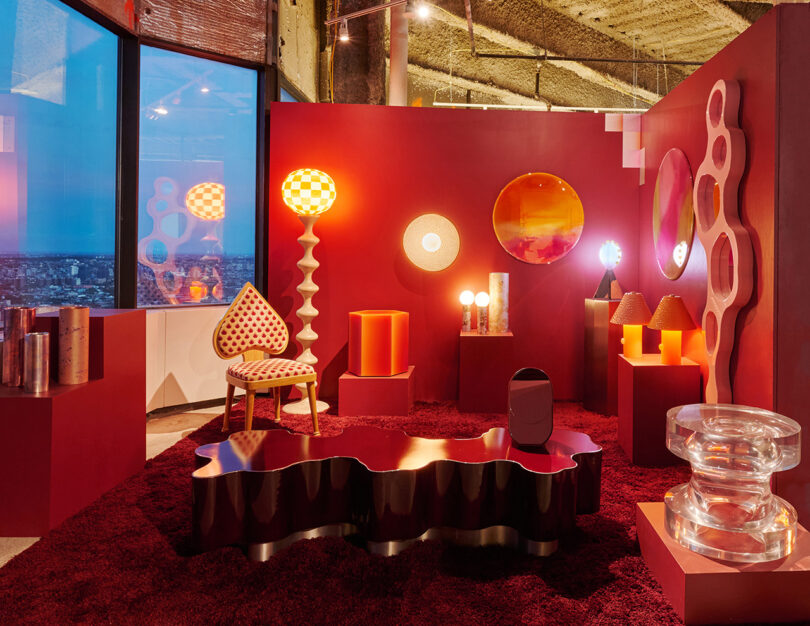 The modern room contains bold red walls and decor, mirrors emphasis, geometric lamps, scene furniture and sculptural items, with large windows overlooking the city.