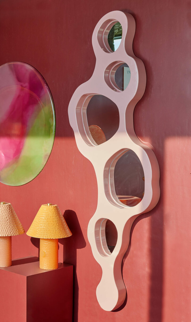 A sculptural wall mirror with multiple irregularly shaped sections is mounted on a red wall near two yellow lamps and a round, reflective artwork.