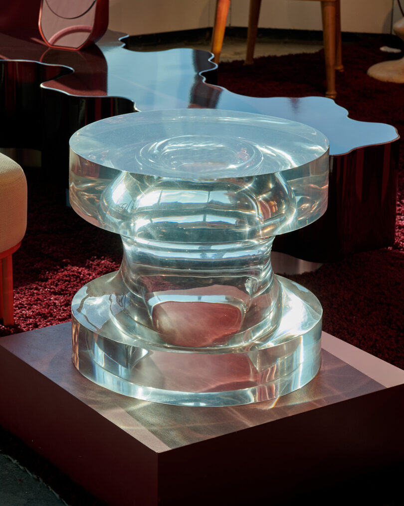 A transparent, sculptural side table made of clear material sits on a reflective surface in a modern, stylish room with red carpeting and contemporary furniture.