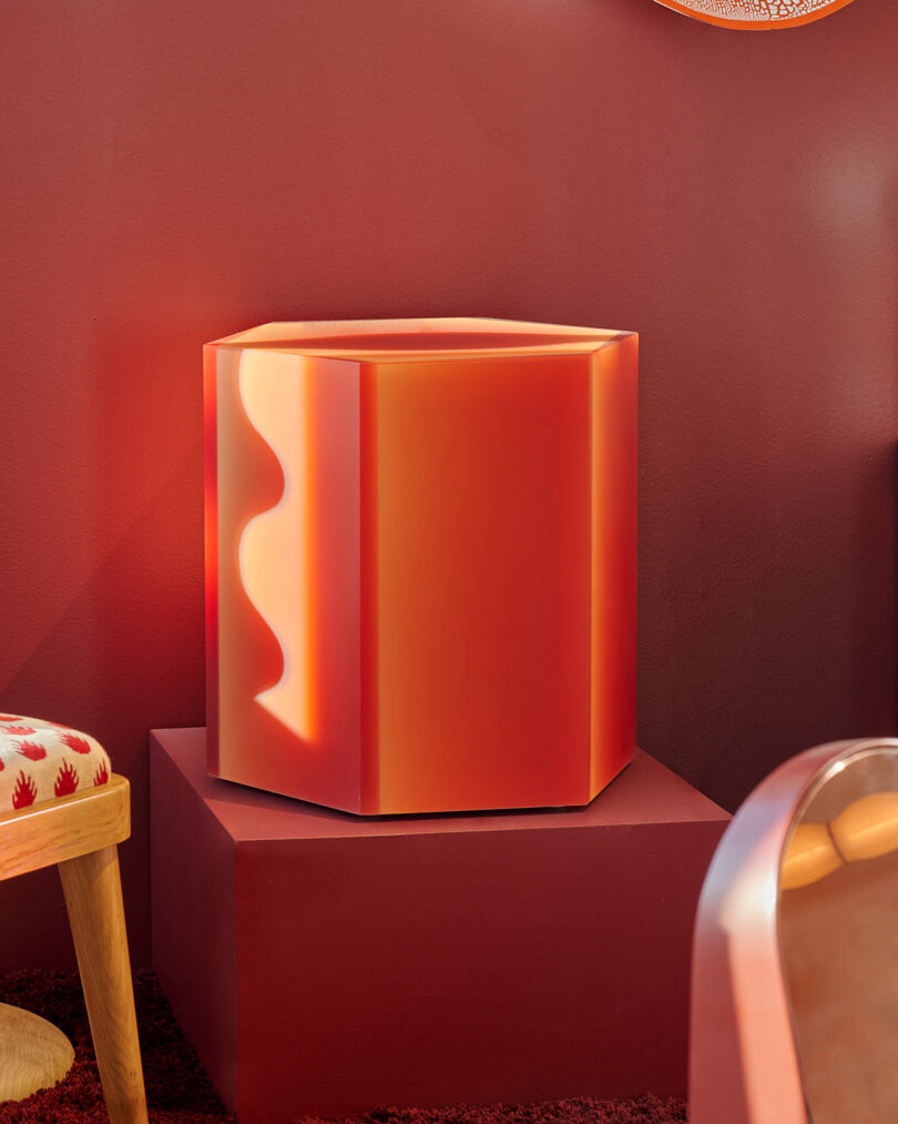 A geometric, orange-red side table with wavy pattern stands on a matching pedestal against a red wall, next to a wooden chair with a patterned cushion.