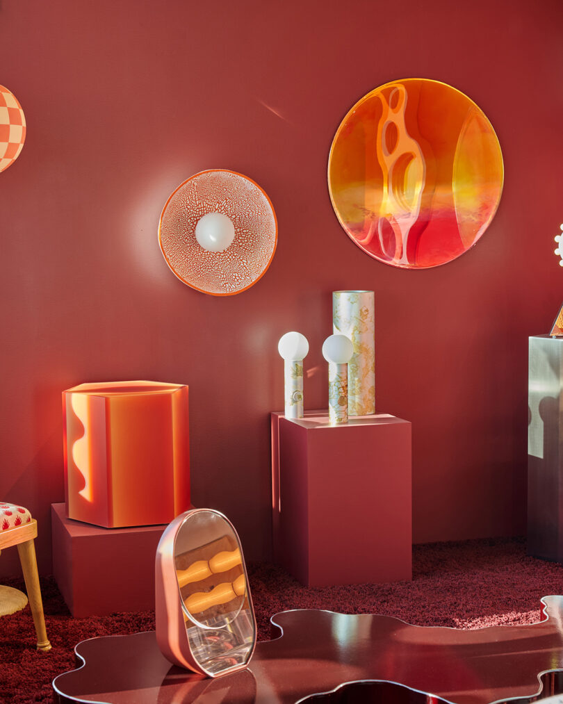 A modern room with red walls, geometric decor pieces, sculptural lamps, circular wall art, and reflective surfaces in warm hues of orange and gold.
