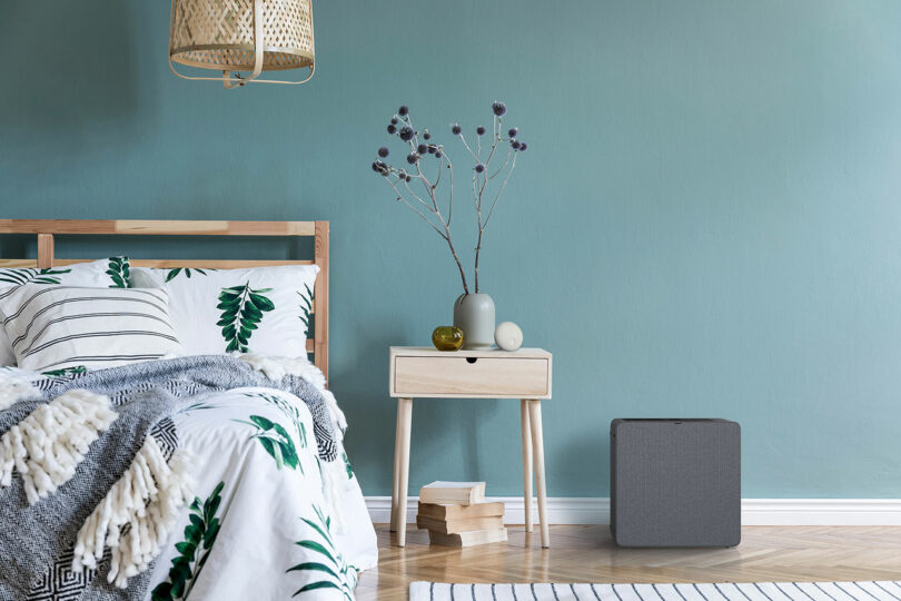El dormitorio tiene una cama de madera con un jarrón y libros en la mesita de noche, un purificador de aire gris en el suelo y paredes verde azulado.