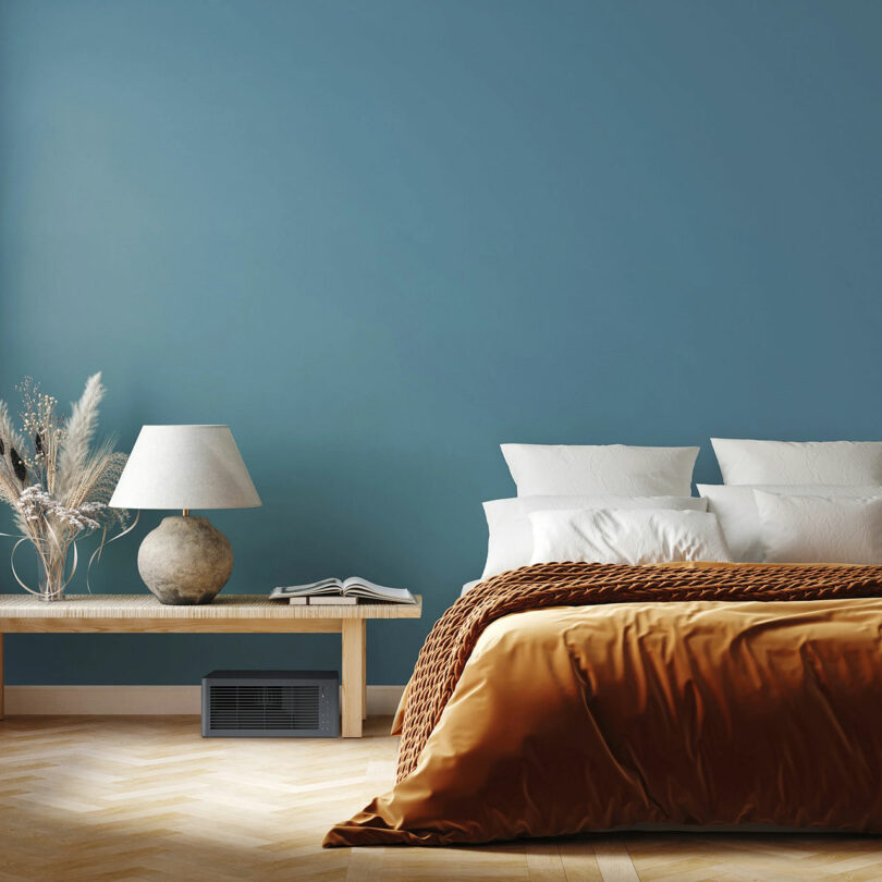 A modern bedroom with a blue wall, a bed with white pillows and a brown blanket, a wooden bench with a lamp, decorative vase, a book, and a vent below.