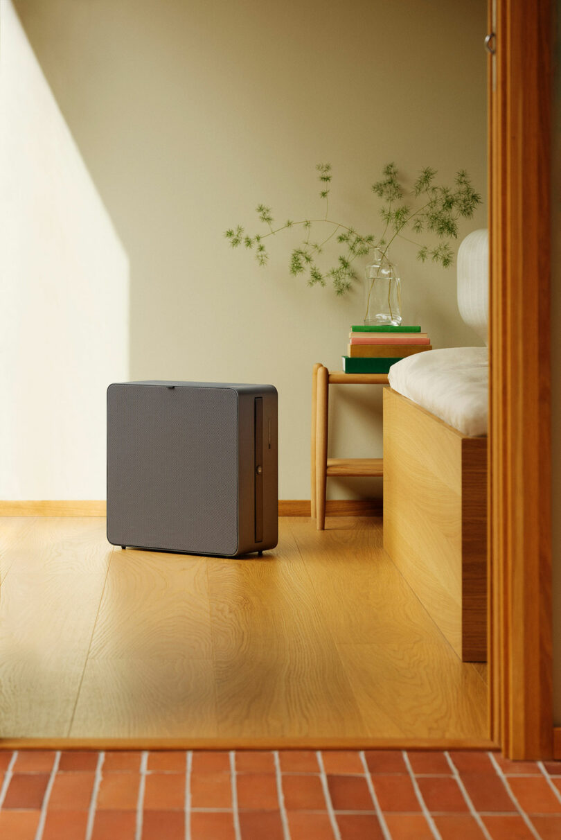 A minimalist room with a gray rectangular air purifier on a wooden floor, next to a bench, a side table with books, and a glass pitcher with water and green sprigs.