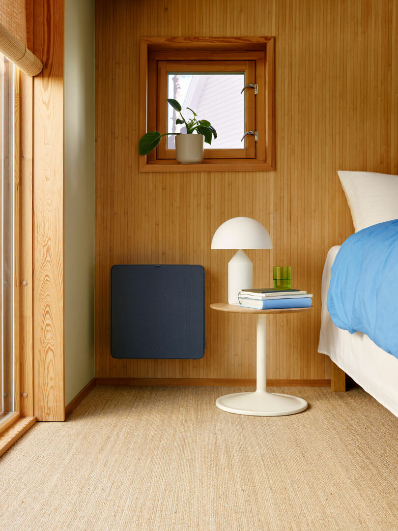 Minimalist bedroom corner with wood-paneled walls, small window, potted plant, round white side table holding a white lamp, green glass, and books beside a bed with blue bedding.