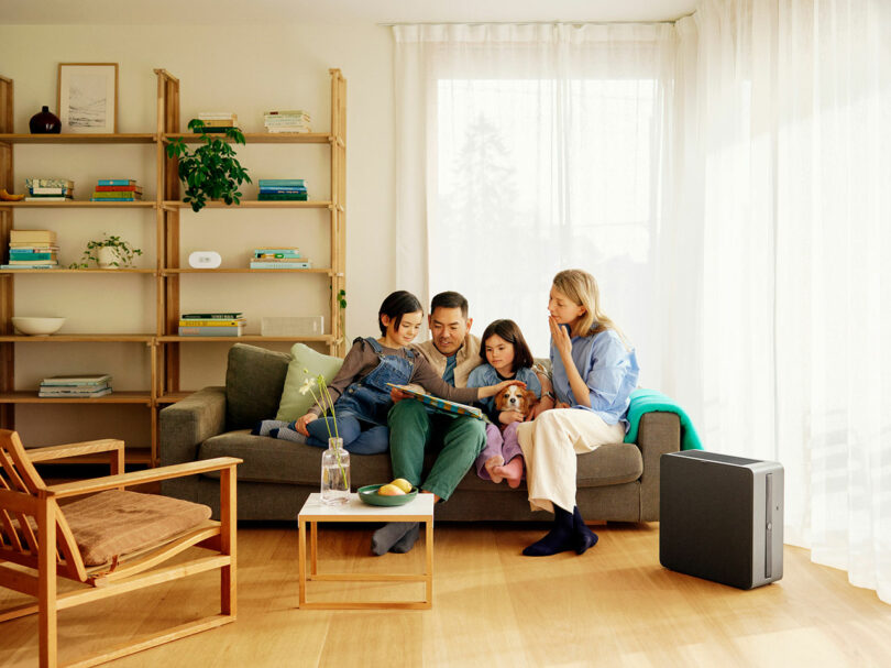 A family of four sits on a couch in a living room, reading a book together. There are shelves with plants and books in the background and a coffee table with a vase in front of them.