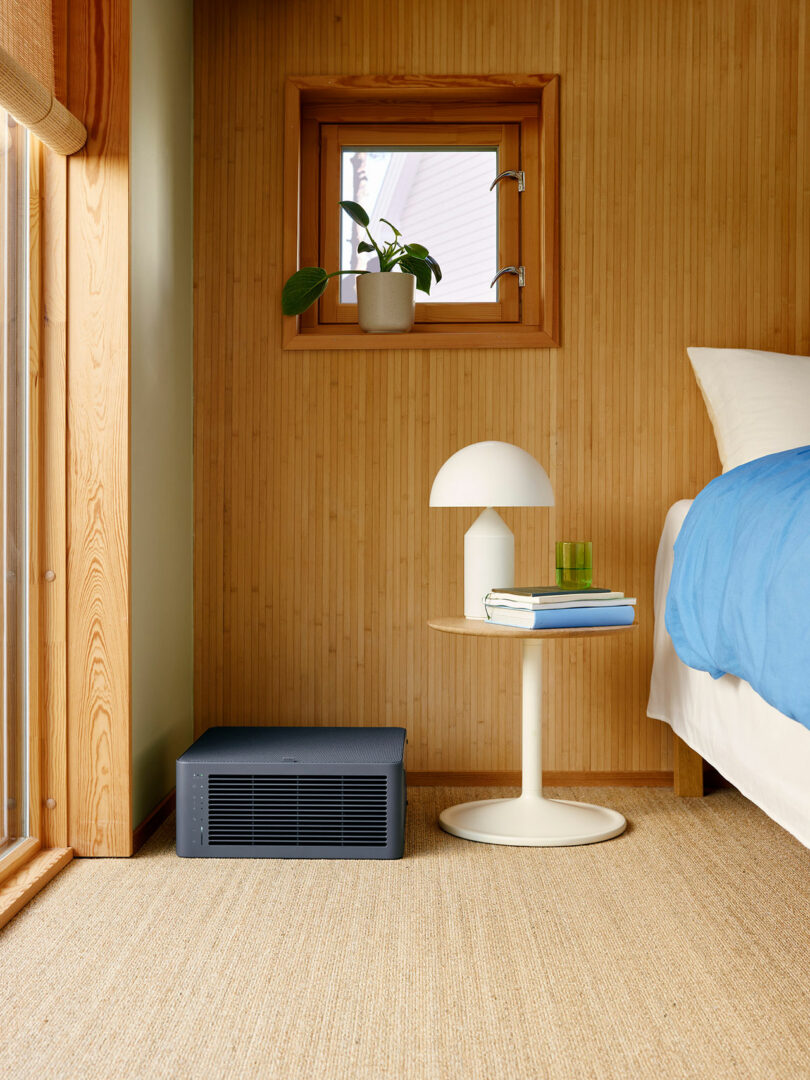 A small black air purifier sits on the floor next to a round white bedside table with a lamp, books, and a glass, in a wood-paneled bedroom with a small window and potted plant.