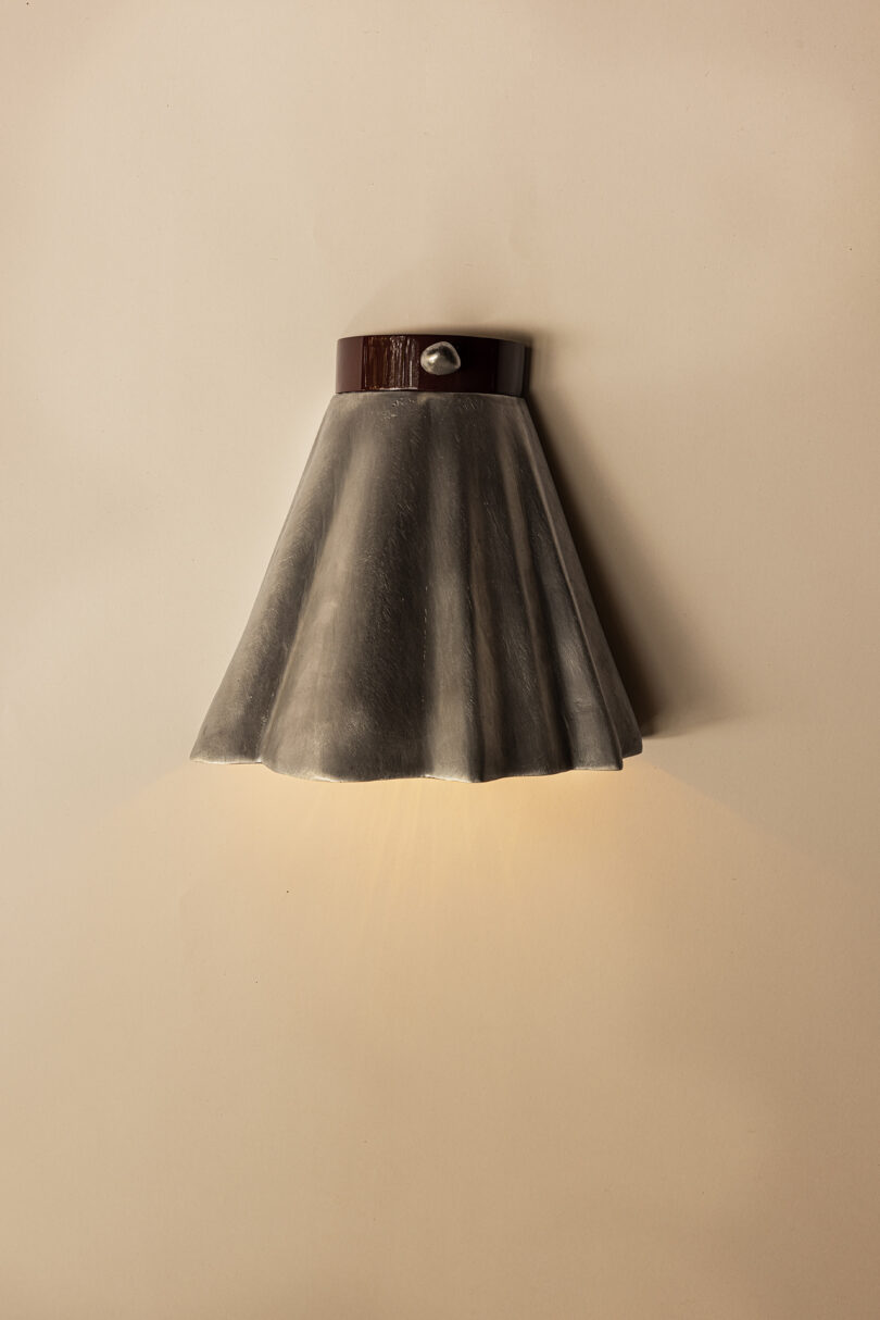 Wall-mounted light fixture with a skirt-shaped, dark lampshade and a wooden accent at the top while casting a soft glow on a beige wall.