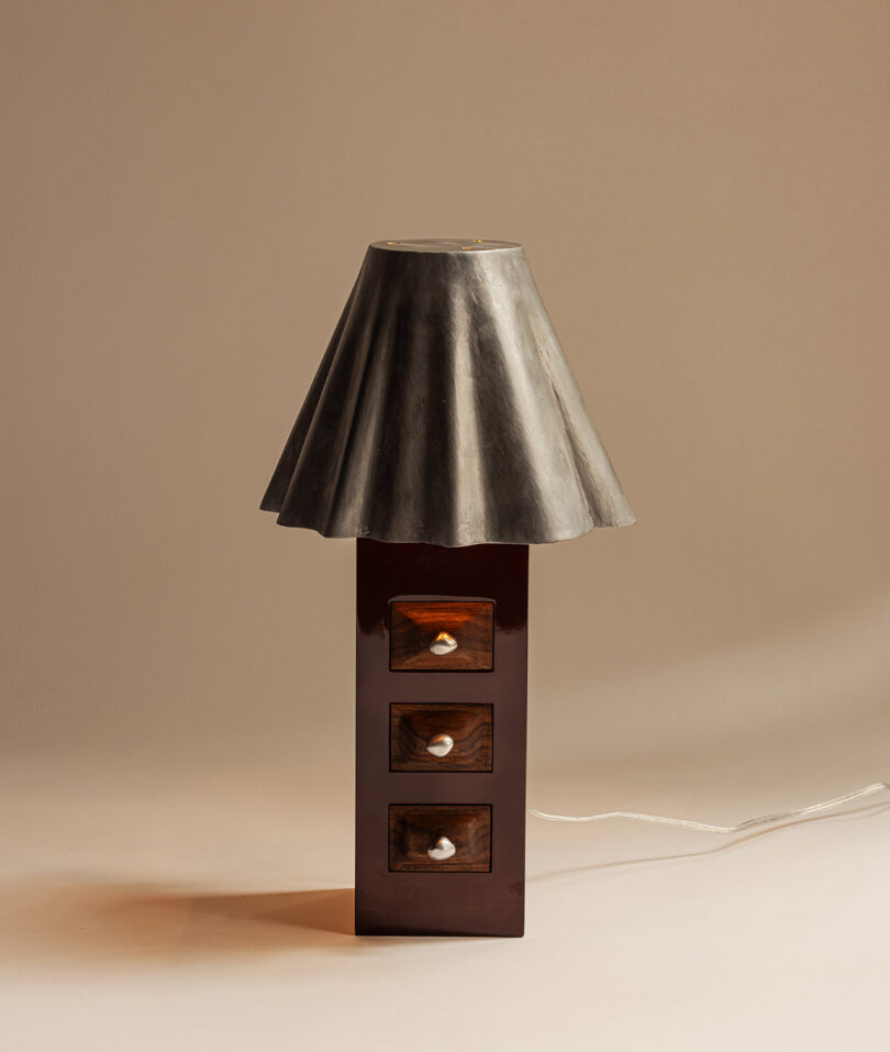 A table lamp with a metallic, flared lampshade and a base resembling a three-drawer wooden cabinet set elegantly against a beige background.