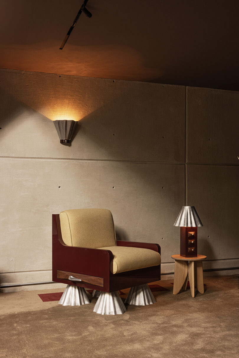 A modern armchair with metallic cone feet sits next to a small wooden table adorned with a silver lamp against a concrete wall with a matching wall sconce.
