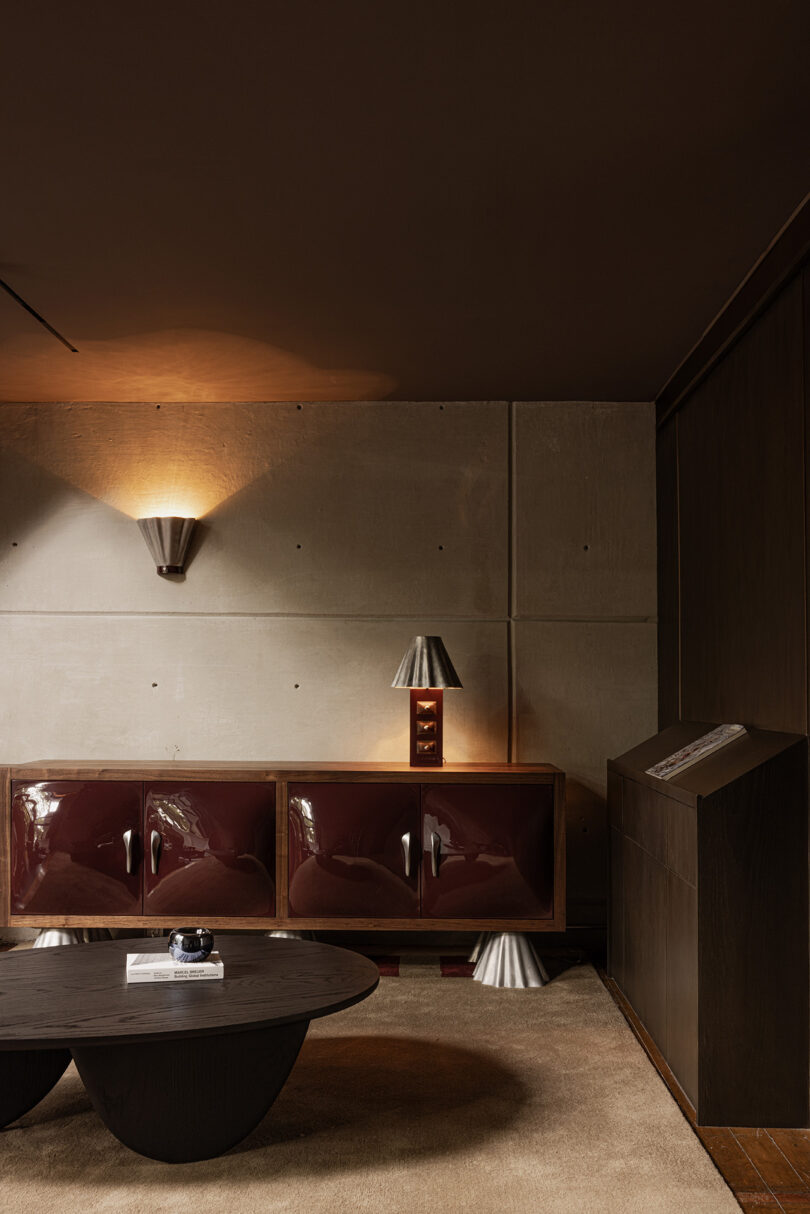 Modern living room with concrete walls, dark brown ceiling, glossy burgundy cabinets, metallic table lamp, stylish wall sconce, and a rounded black coffee table on a beige rug—designed with all the bells & whistles for contemporary comfort.