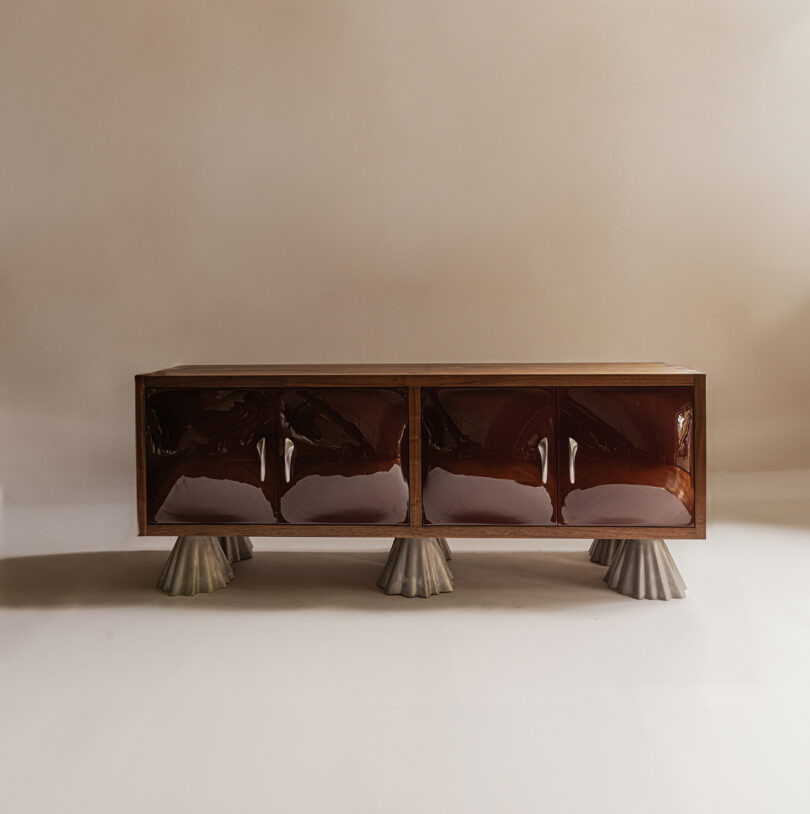 A low, rectangular wooden cabinet with glossy, dark brown doors and four sculptural, cone-shaped gray legs on a neutral background.
