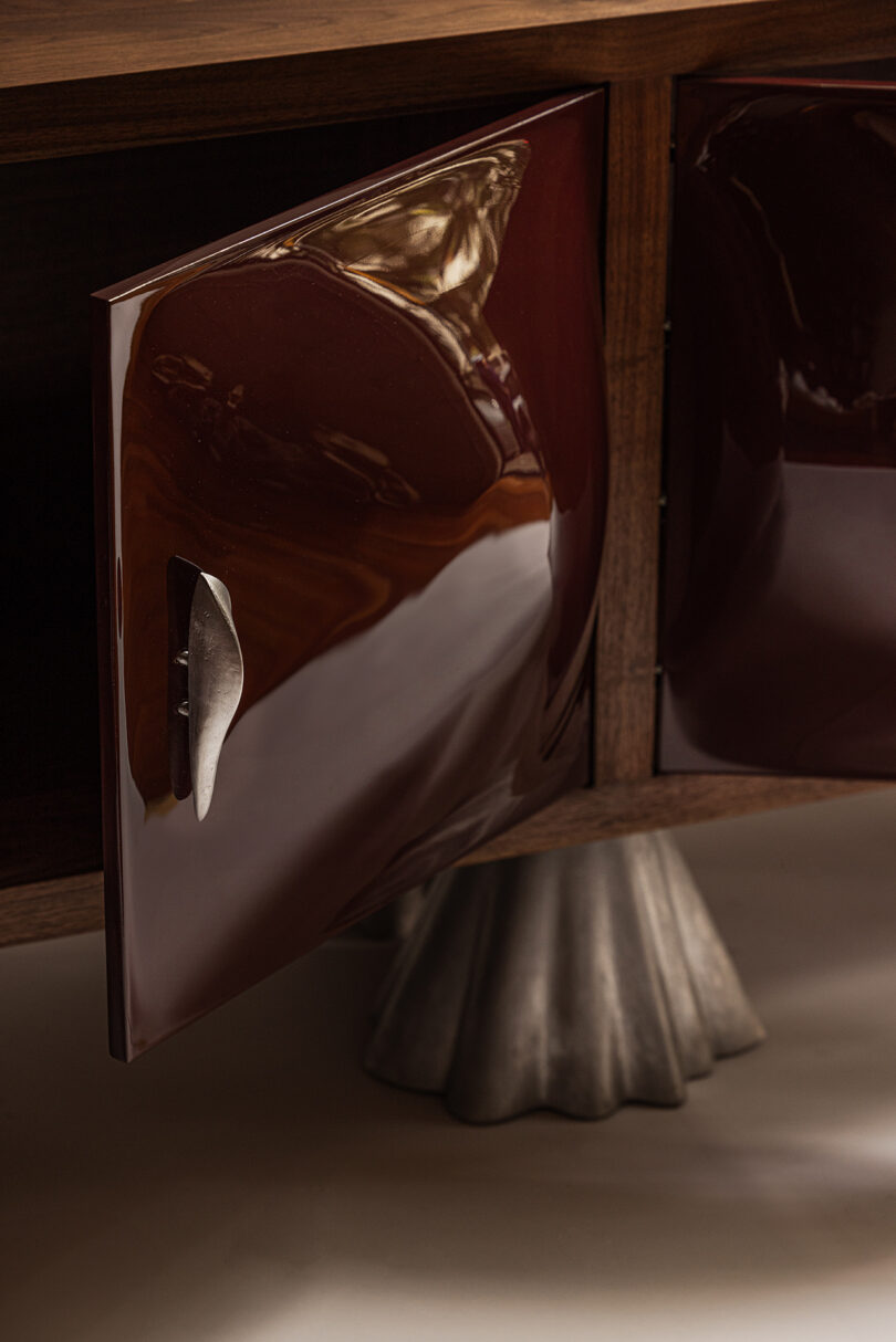 Close-up of a wooden cabinet with a glossy, dark brown door partially open, featuring a sculpted metallic handle and a textured metallic base.