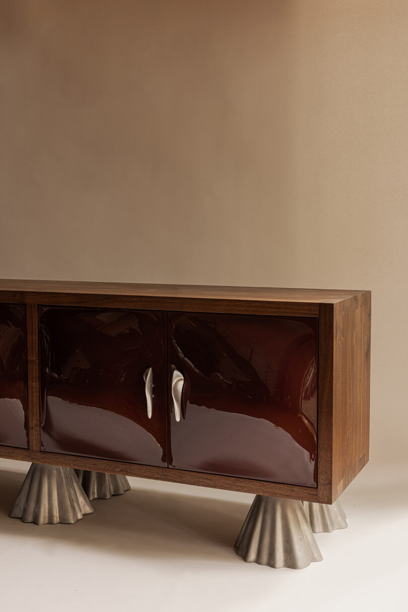 A wooden cabinet with glossy dark brown doors, metallic silver handles, and four sculpted silver feet resembling shells, set elegantly against a neutral background.