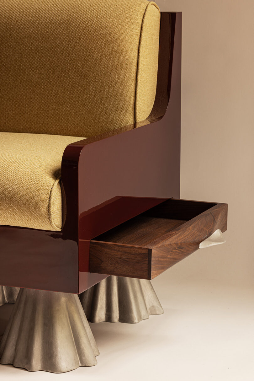 A modern armchair with yellow upholstery, glossy maroon sides, metallic flared legs, and a wooden side drawer partially pulled out.