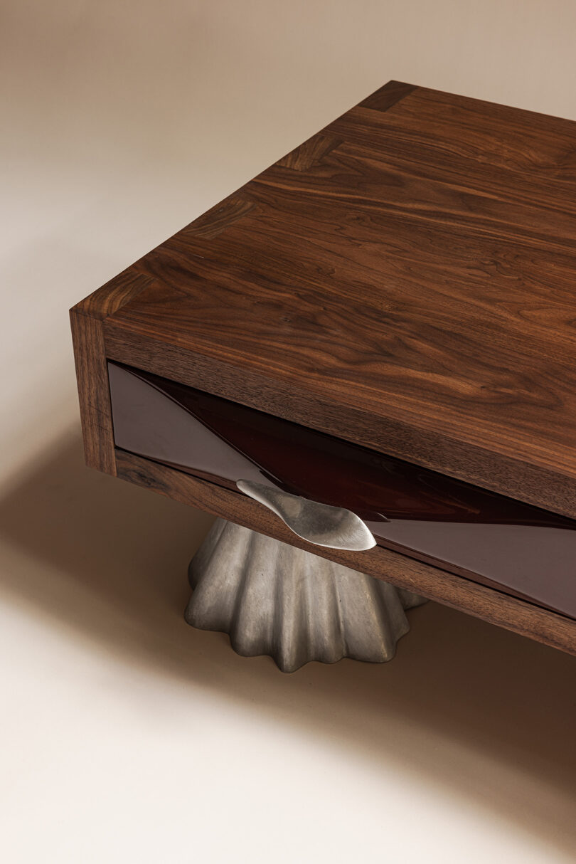 Rectangular wooden table with a glossy maroon accent drawer and a sculpted metallic leg resembling draped cloth, featuring subtle Bells & Whistles for a touch of sophistication.