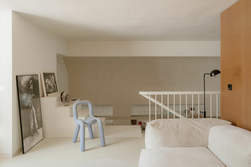 Minimalist loft interior with neutral tones, featuring a white sofa, a light blue modern chair, framed artwork on the floor, and a railing overlooking the lower level.