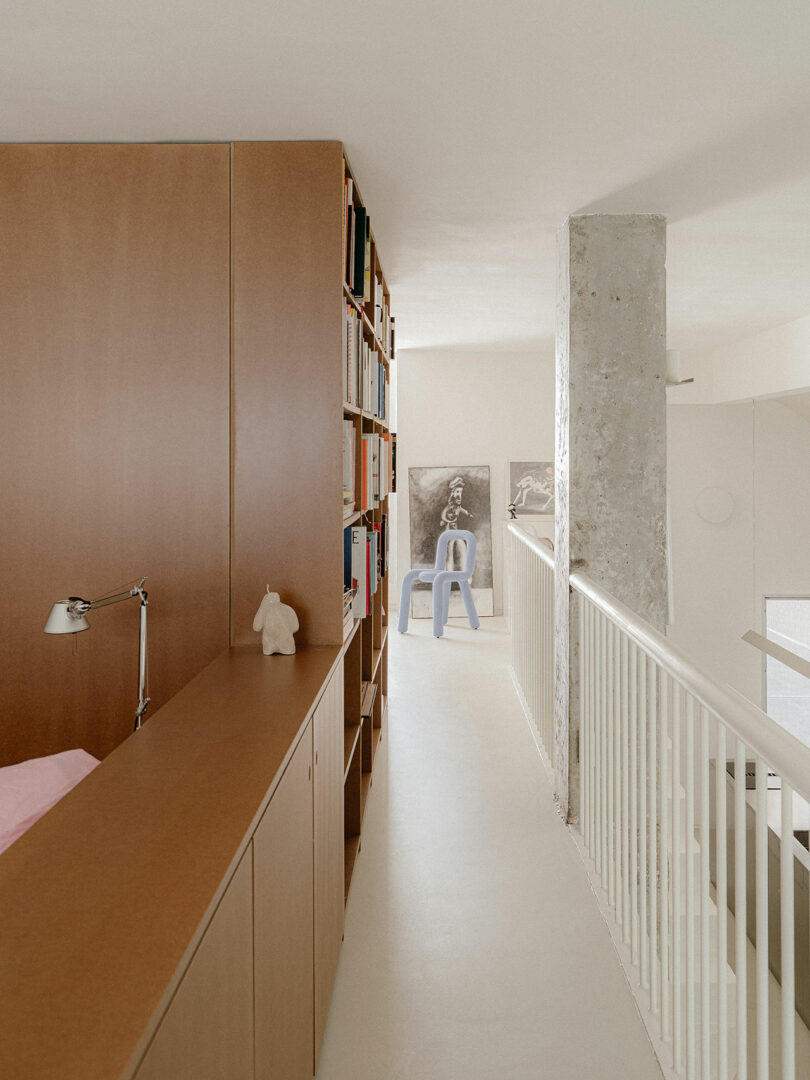 A hallway with built-in wooden shelves holding books, a desk lamp, and a small sculpture; at the end are two monochrome artworks and a light blue chair.