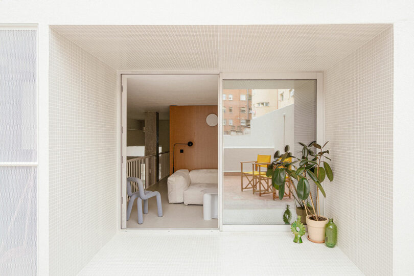 A modern balcony with a sliding glass door opens to a minimalist living space with white walls, a sofa, a chair, potted plants, and an outdoor table with yellow chairs.