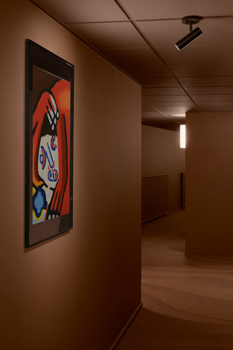 A framed abstract painting with a red background hangs on a brown wall in a dimly lit hallway with a spotlight above.