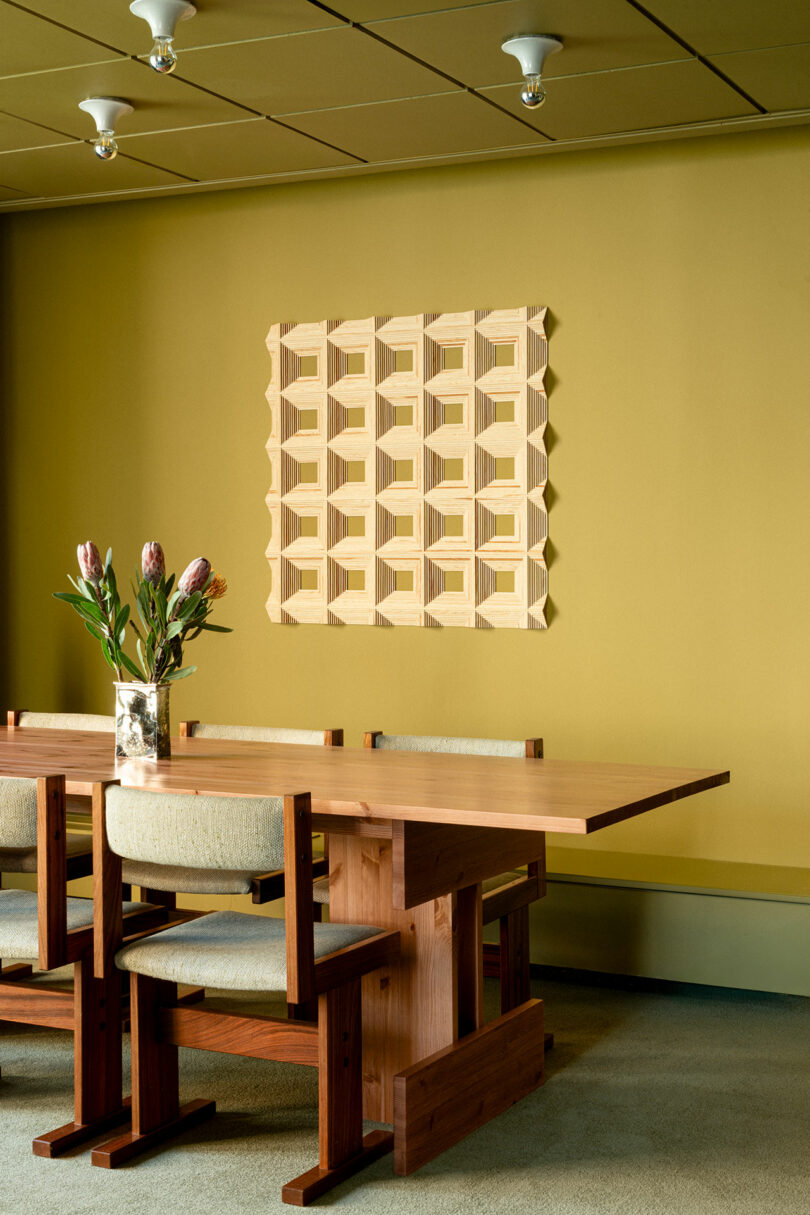 A wooden dining table with four chairs is set against a mustard yellow wall, decorated with a geometric wooden art piece; a vase with flowers sits on the table.