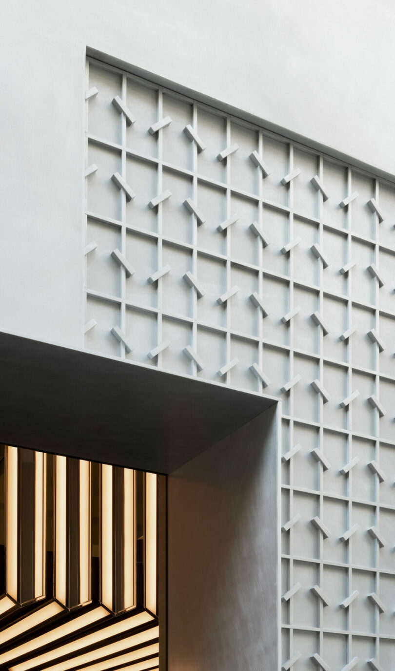 A geometric wall with a white grid pattern is positioned above a modern structure featuring illuminated horizontal and diagonal light strips.
