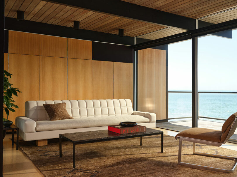 Modern living room with wood panel walls, a white tufted sofa, a marble coffee table, a tan accent chair, and floor-to-ceiling windows overlooking the ocean.