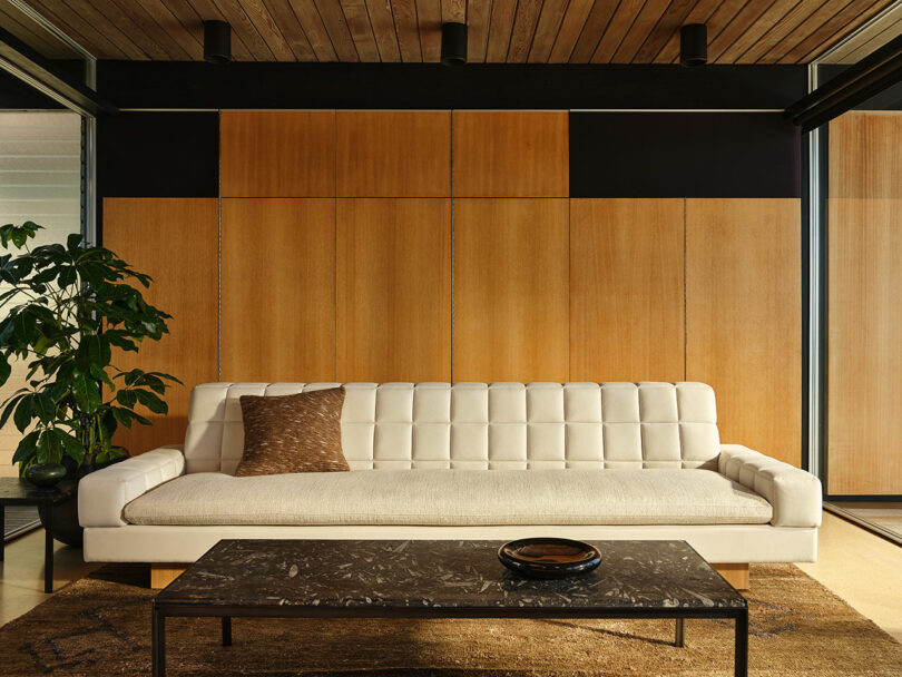 Modern living room with a white tufted sofa, a brown pillow, a marble coffee table, a potted plant, and wood panel walls.