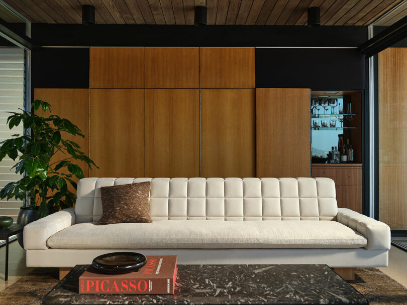A modern living room with a white tufted sofa, a brown cushion, a black marble coffee table with books and a tray, and wood-paneled walls with a potted plant on the left.
