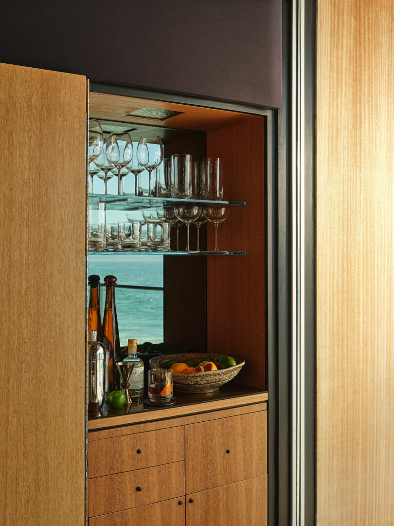 A wooden cabinet with glass shelves holding wine glasses, bottles, and a fruit basket, positioned next to a window overlooking the sea.