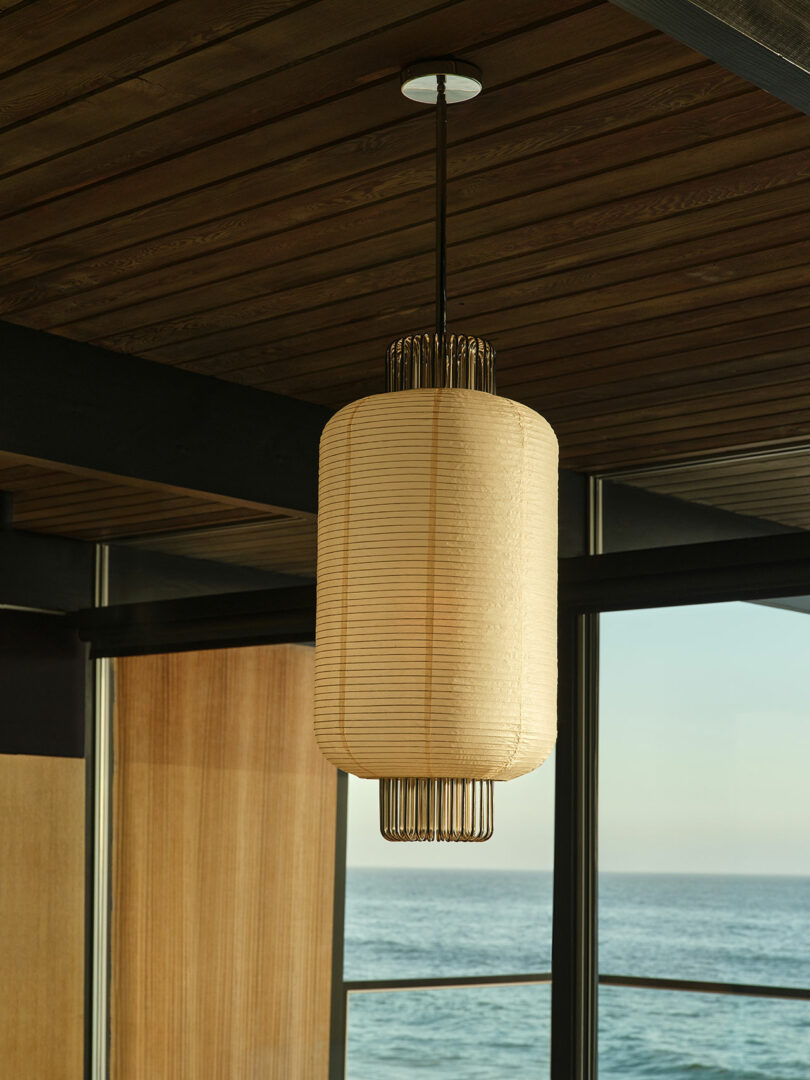 A cylindrical pendant light with a ribbed, beige shade hangs from a wooden ceiling in a modern room with large windows overlooking the ocean.