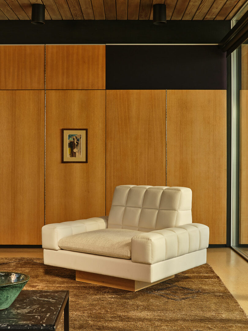 A modern living room with a large white upholstered armchair on a brown rug, wooden paneled walls, a small framed artwork, and a green decorative bowl on a table.