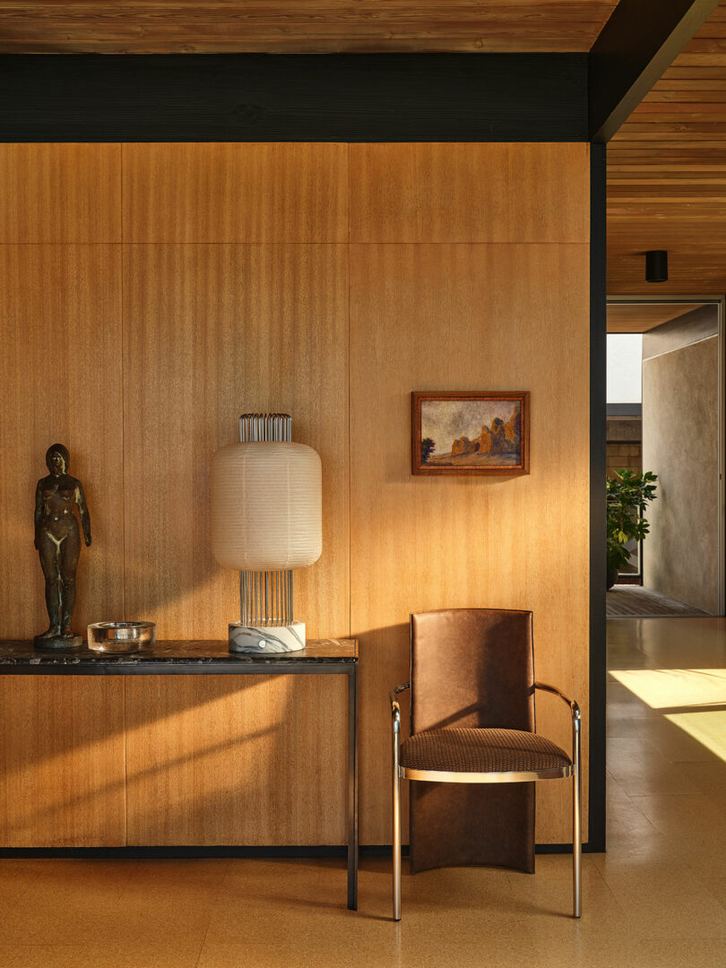 A wood-paneled wall with a console table holding a sculpture, a lamp, and a bowl, a framed artwork above, and a brown leather chair beside it in a sunlit room.