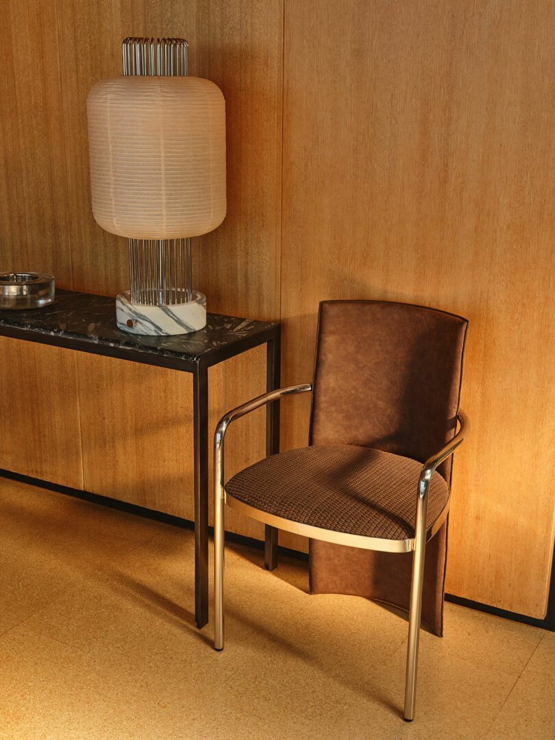 A brown cushioned chair with metal arms sits next to a black marble-top table holding a large modern lamp, against a wooden wall.