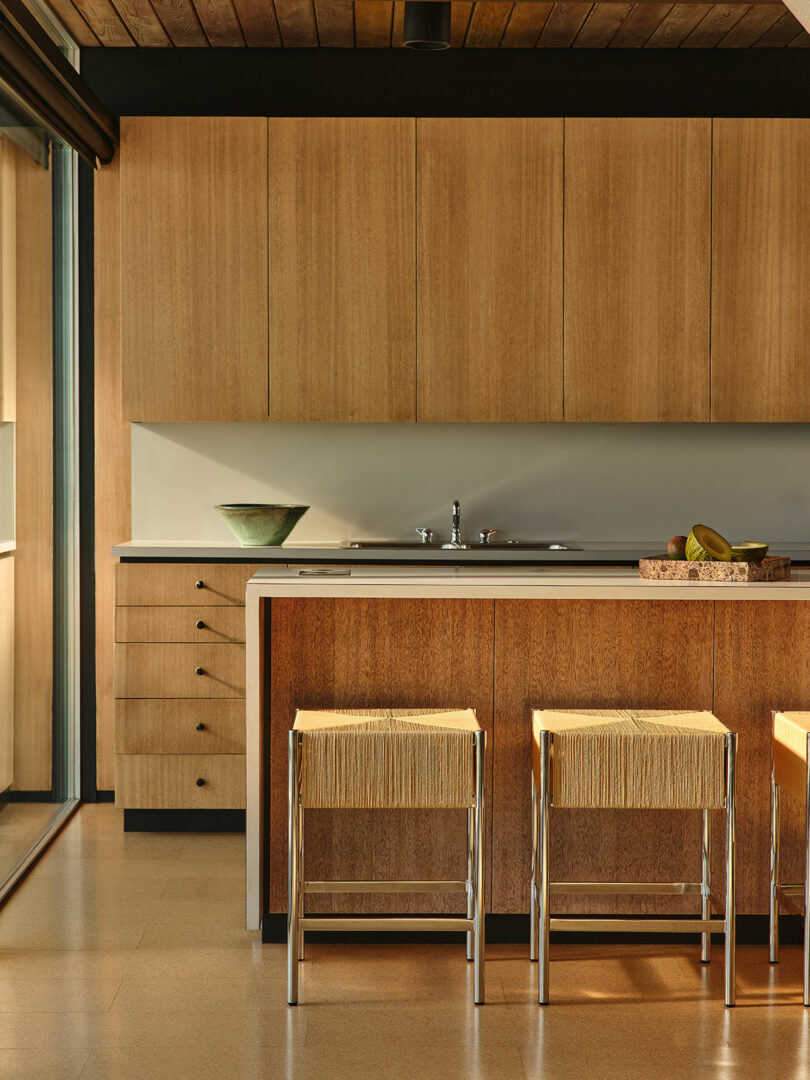 Modern kitchen with wood cabinets, a white countertop, three bar stools with woven seats, a bowl, and a basket with fruit on the counter.