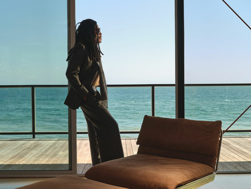 Person in a suit stands indoors by large windows, looking out at the ocean and wooden deck, channeling a calm, confident vibe reminiscent of Lenny Kravitz on a clear day.