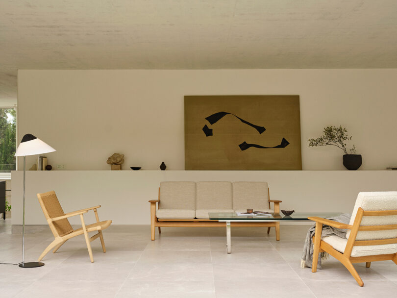 A minimalist living room with light wooden furniture, a glass coffee table, abstract wall art, and neutral decor on a wide shelf against a white wall.